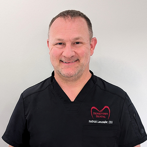 Male dental professional in black scrubs smiling at camera in clinical setting.