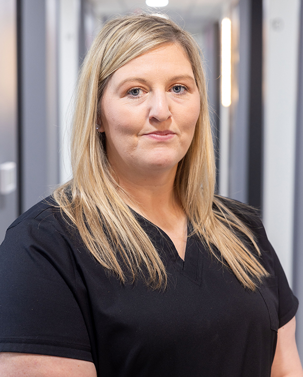 Professional dental staff member in black scrubs smiling at camera in clinical setting.