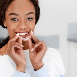 Woman smiling while holding clear dental aligner, modern dental office setting background.