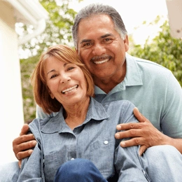 Happy middle-aged couple smiling outdoors in casual clothing, embracing warmly in front of house.