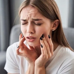 Woman touching her cheek in pain, appearing to have a toothache or dental discomfort indoors.